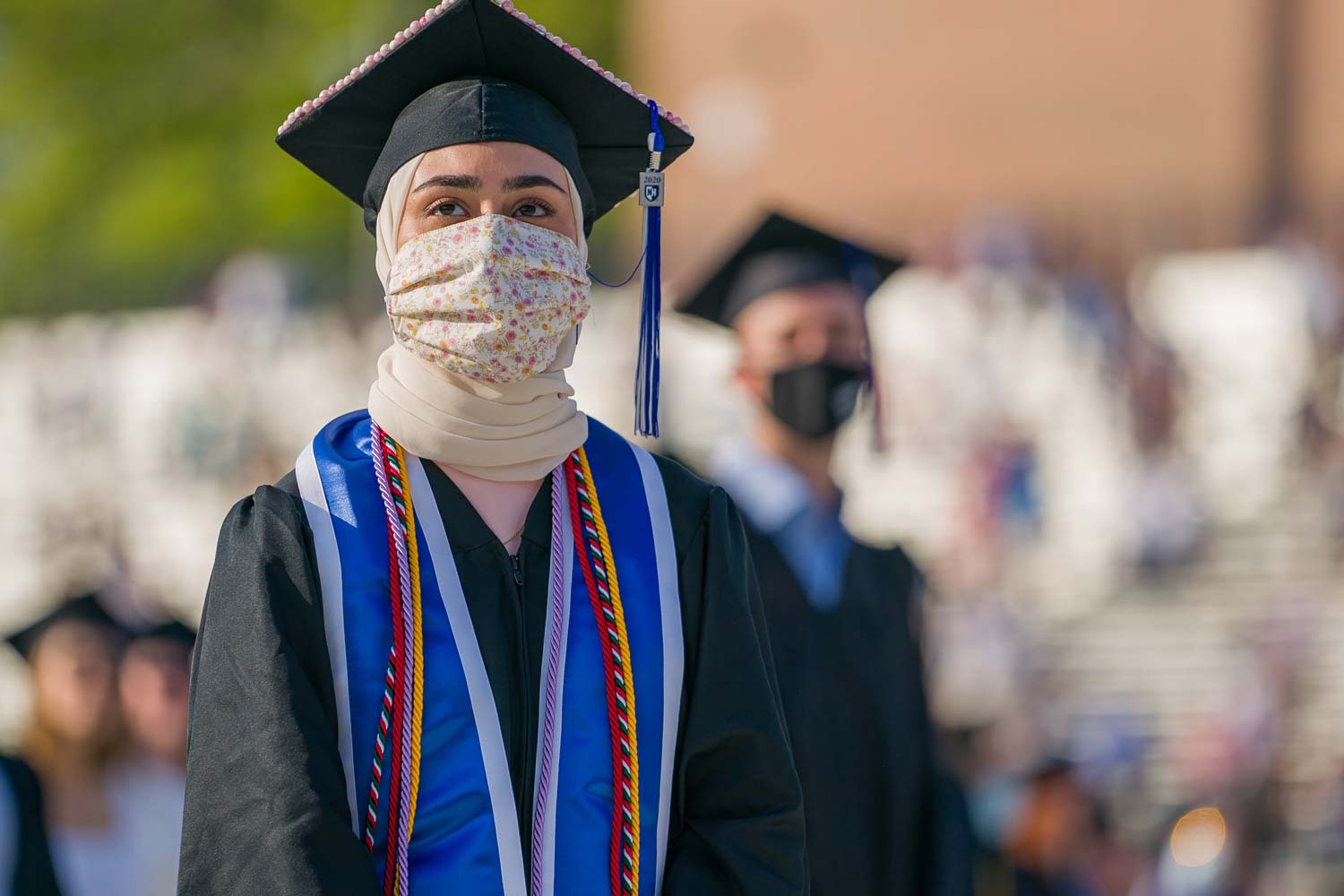 A student wearing a cap, gown, and floral head covering watches the ceremony with their hands held in respect