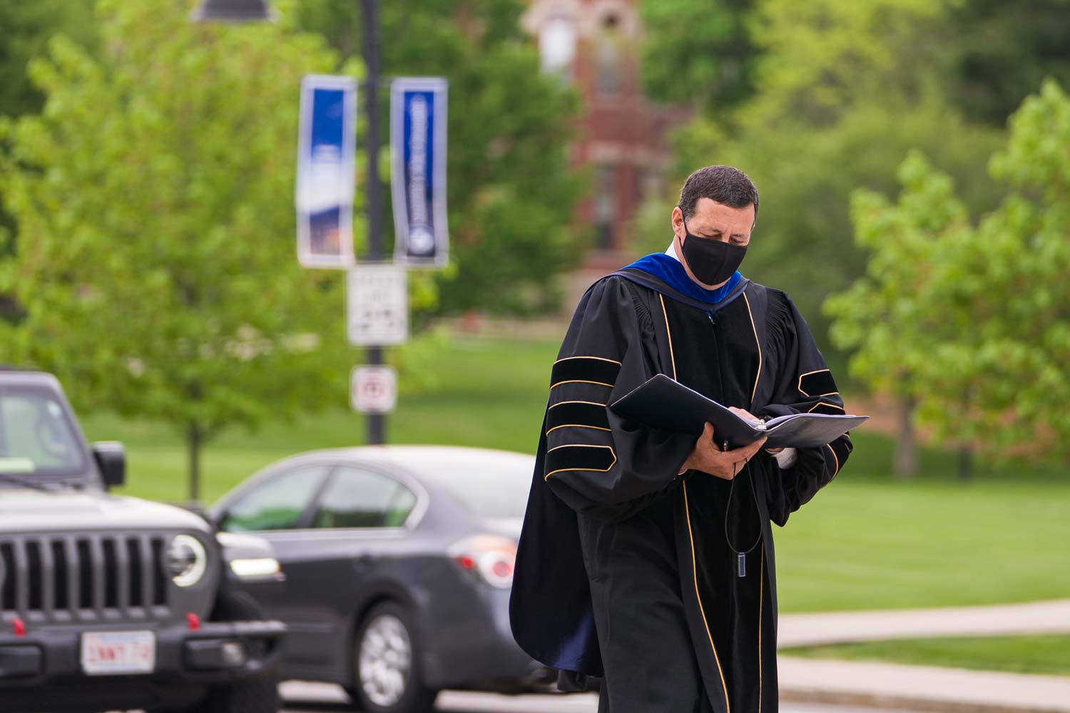 A professor in a gown and mask reading from a binder while walking on the sidewalk