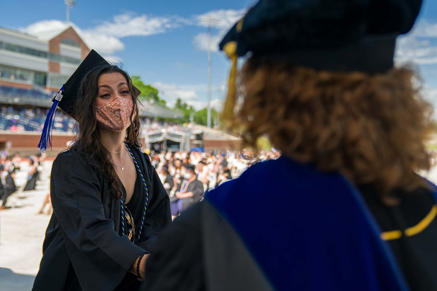 Student wearing a mask, cap, and gown shakes a professors hand as she receives her diploma
