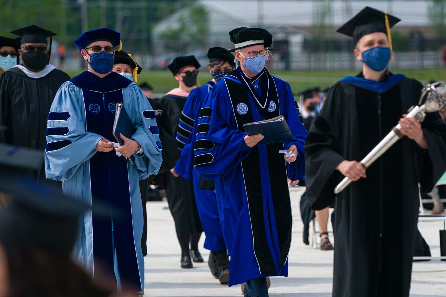 The dean and several other professors and speakers clad in gowns and caps walk through the center aisle at the graduation ceremony