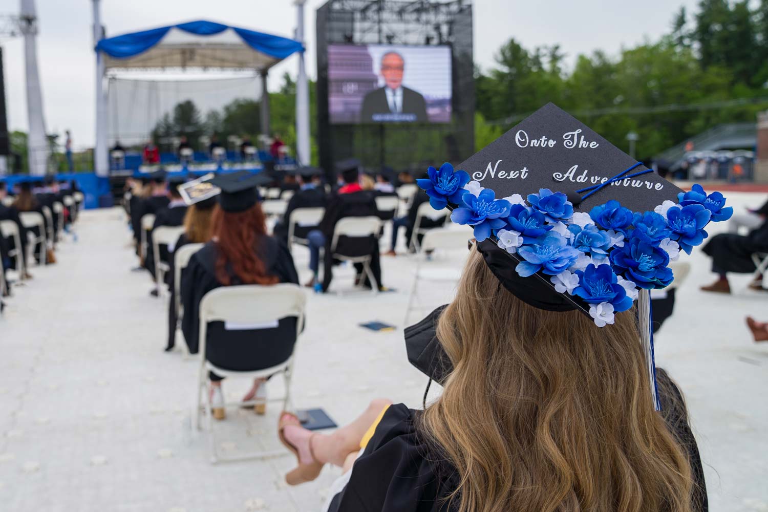 Student watching the ceremony and wearing a graduation cap that reads "onto the next adventure" in curly cursive lettering with blue and white flowers collaged at the bottom