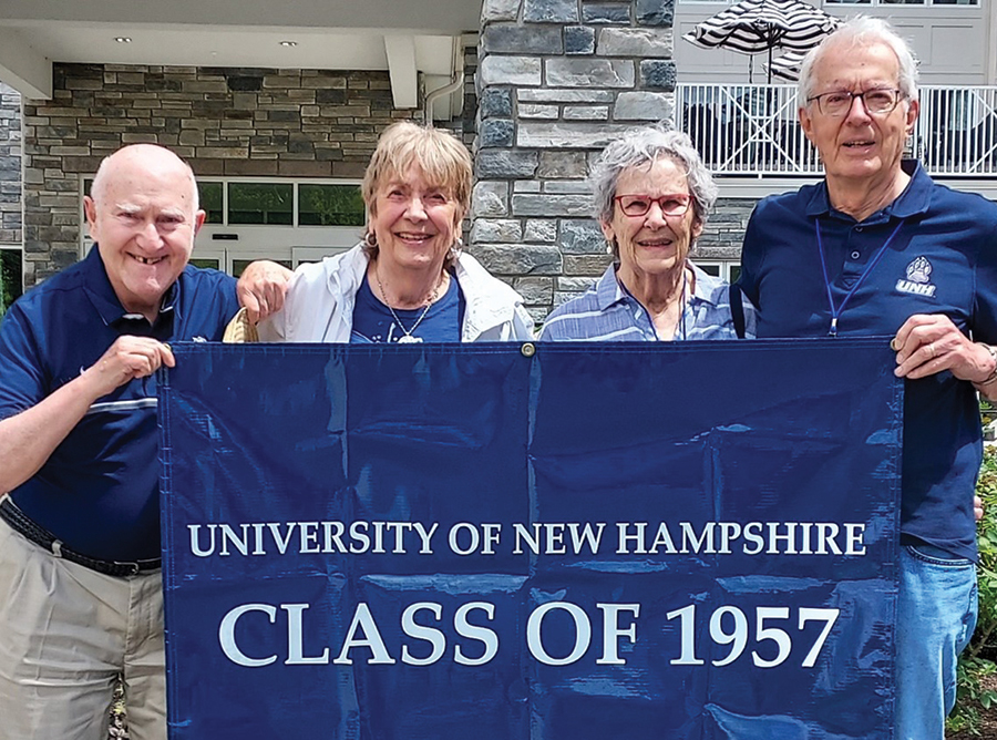 A landscape photograph of Michael Gordon '57, Nancy Glowacki '57, Carly '57 and Jim Hellen '57 celebrating Homecoming together outside smiling and posing for a picture together in their UNH spirit attire as all four individuals are holding a University of New Hampshire Class of 1957 blue flag banner