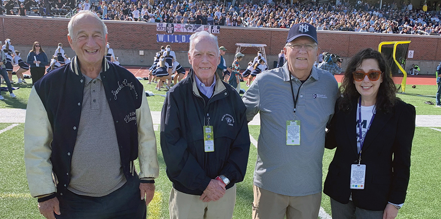 A landscape photograph of Chuck Glenn '63, Bob Towse '63, Jim Twombly '63 and UNH Athletic Director Allison Rich smiling and posing for a picture together in their UNH spirit attire on the field on a bright sunny day at the Elon game in recognition of the 1962 undefeated UNH football team