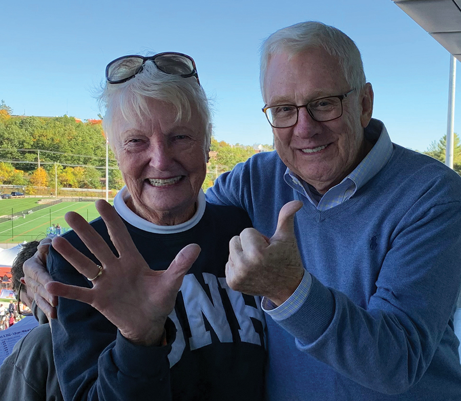 A portrait photograph of Carroll Winch ’63 and Cheryl Dickson ’63 smiling and posing for a picture together holding up six fingers in their UNH spirit attire outside on a bright sunny day to mark the six decades since graduation the class of 1963 will be celebrating at its 60th reunion in June