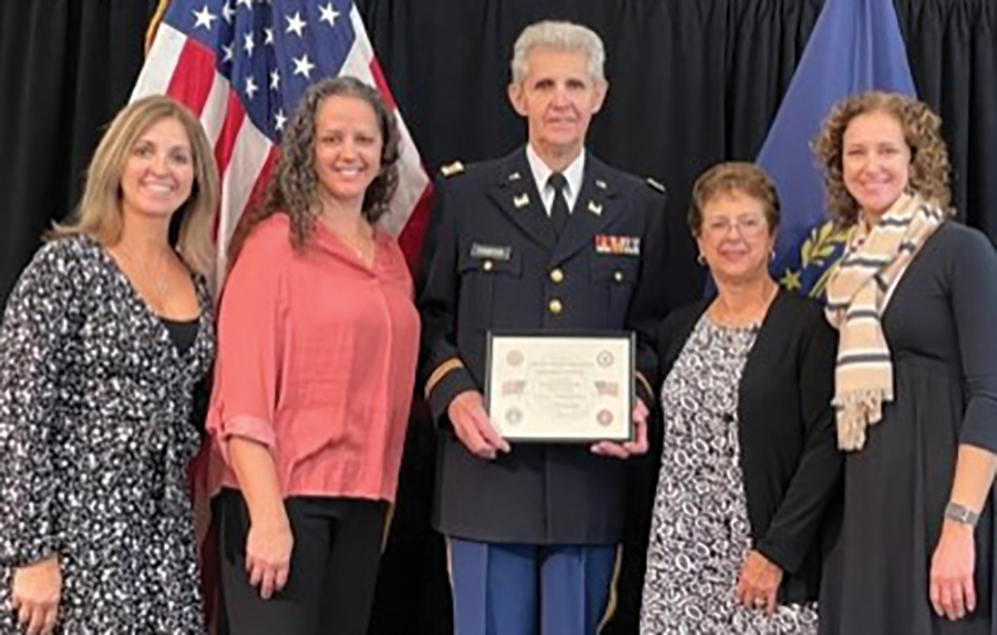 A landscape photograph of Lt. Col. Charles W. Thompson (standing in the middle in his military outfit), ret., surrounded by family as he was inducted into the UNH ROTC Hall of Fame back in November. From left are daughters Kristen Thompson Arensberg ’92, Amy Thompson Olsen and Susan Thompson Rafferty all smiling and posing for a picture together standing in front of the United States of America flag