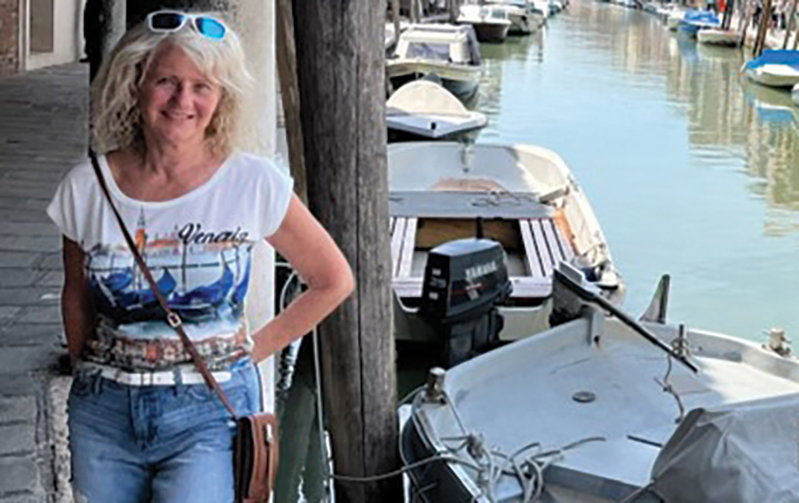A landscape photograph of Berneen Bratt ’69 ’86G traveling and exploring in recent months as she is smiling posing for a picture in a white t-shirt, blue jeans, white sunglasses on top of her head, and a purse around her shoulder standing next to a dock pole nearby some boats and water