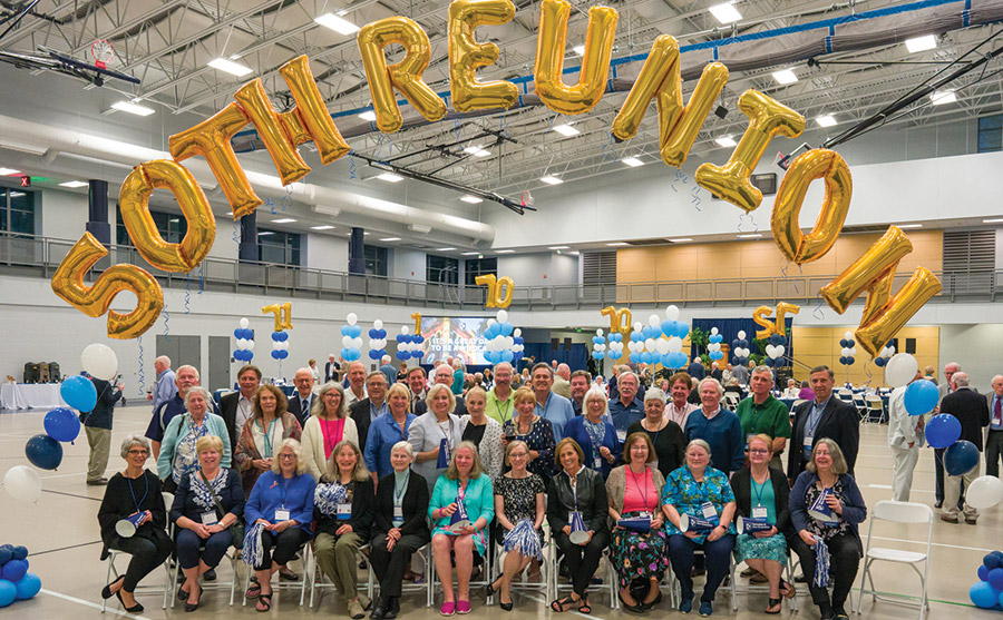 A landscape photograph of classmates from 1970, ’71 and ’72 reunited to celebrate their 50th reunions in June 2022 inside a basketball gym facility posing and smiling for a picture together with a 50th Reunion yellow celebratory ballon arch statement floating above their heads after two years of Covid delays