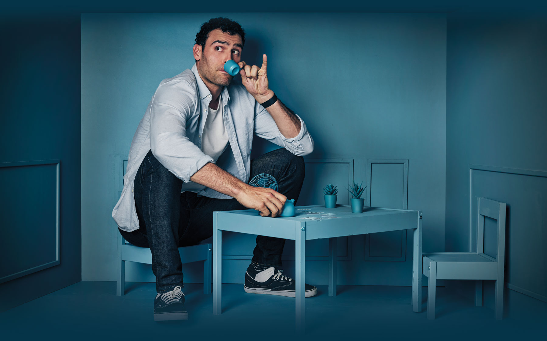 Dane DiLiegro sitting in an all blue room where everything is shrunken down, pretending to sip from a tiny tea cup