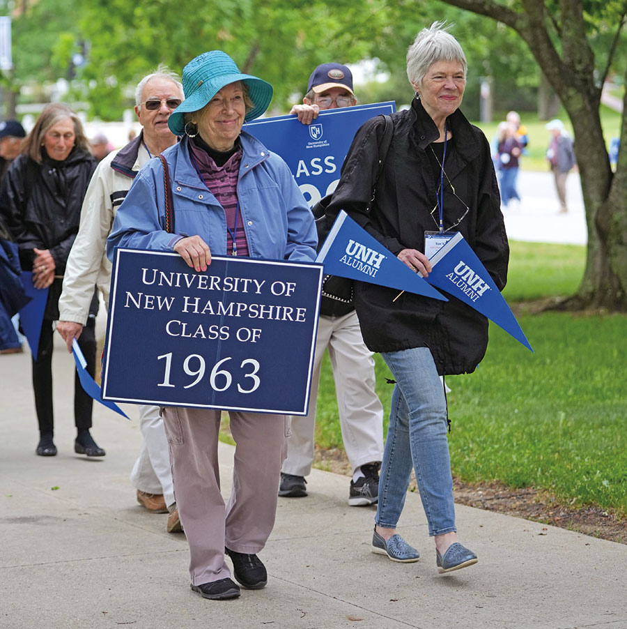 Group of people in background walking on a sidewalk portion of the UNH campus behind two elderly women in foreground grinning as they hold a blue/white University of New Hampshire Class of 1963 yard sign and UNH alumni pennant banners