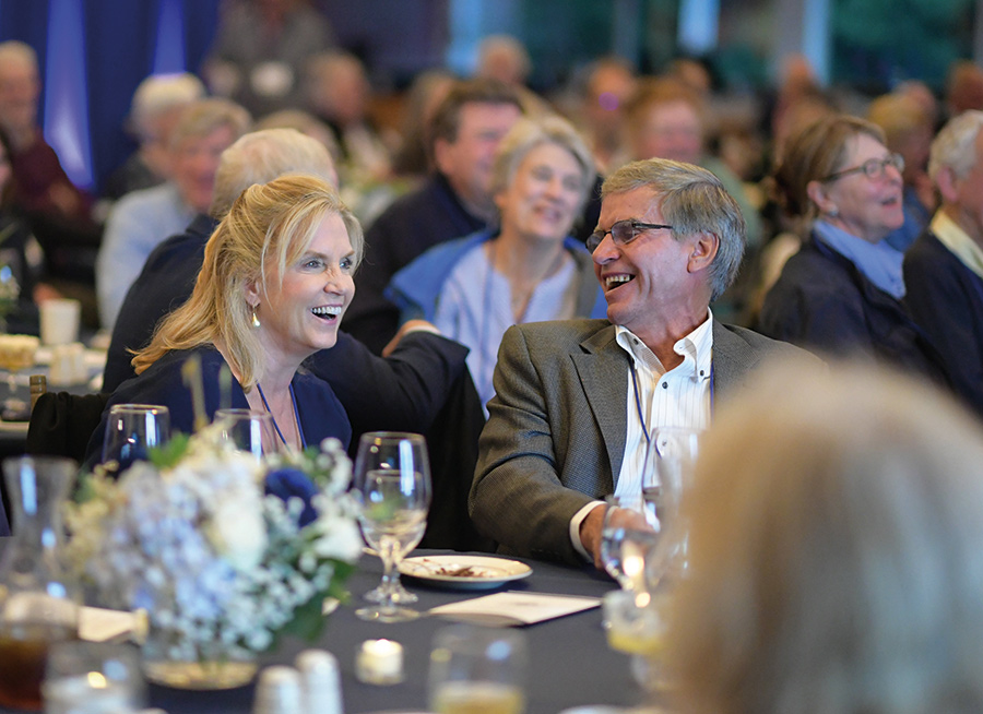 Two individuals (a woman and a man) smile and laugh sitting at a table with wine glasses nearby in the foreground and many other people behind them in the background as they all enjoy a good time together