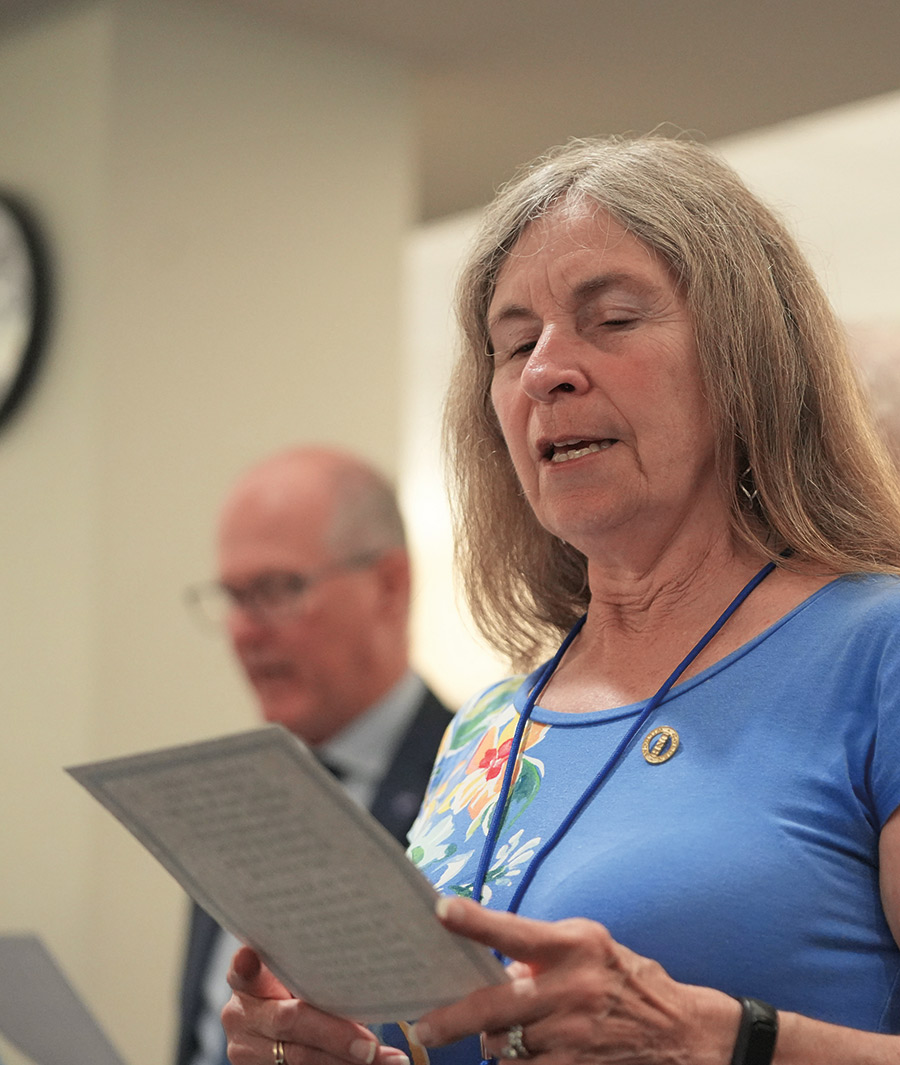 Woman in blue shirt with a floral pattern style holding a piece of paper as she glances down reading information in foreground and another man in background with prescription glasses in suit and tie holds a piece of paper as he glances down reading information