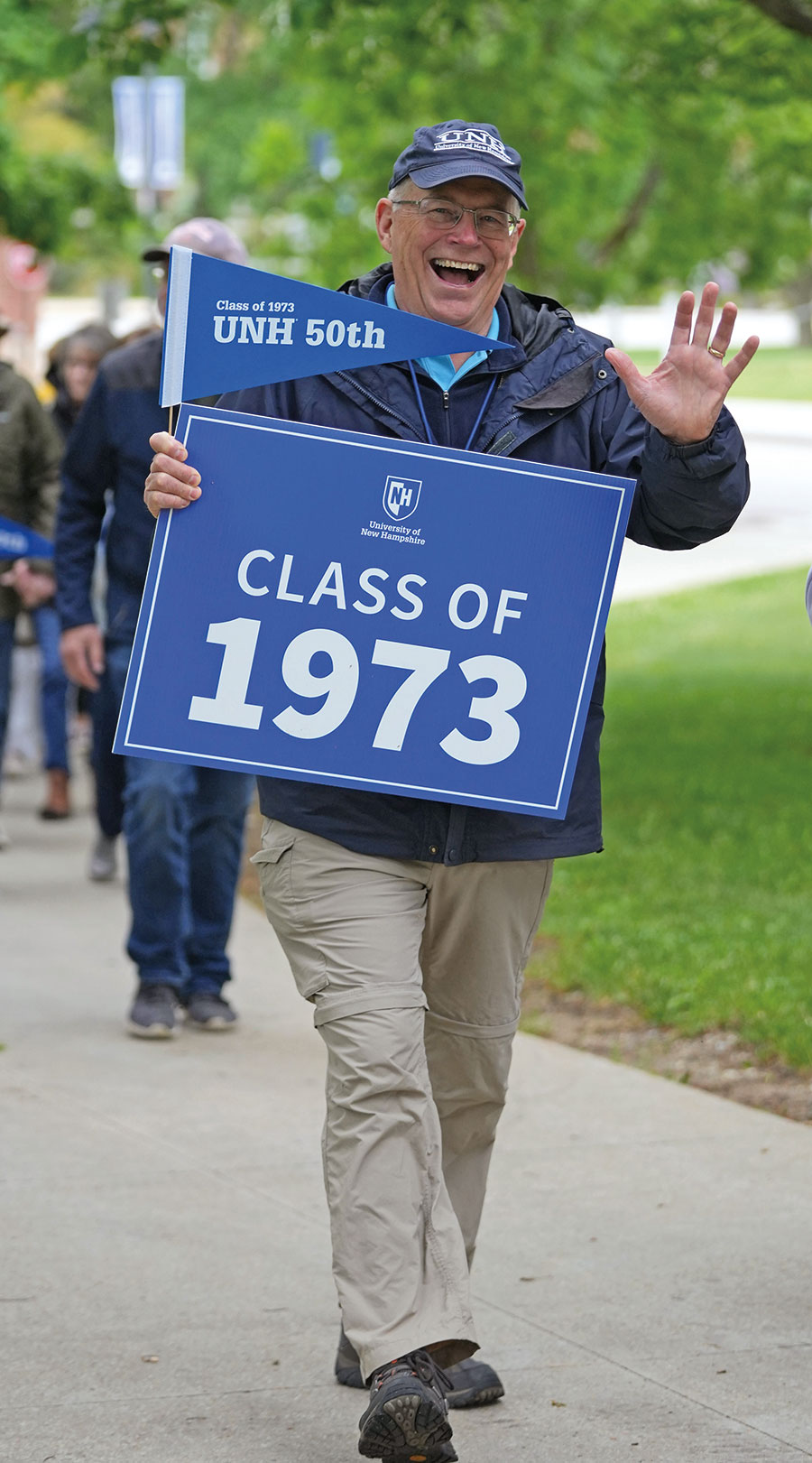 Man in UNH hat and prescription glasses in foreground walking on a sidewalk portion of the UNH campus smiling and laughing as he waves holding a blue/white Class of 1973 UNH 50th pennant banner and UNH Class of 1973 yard sign as there are other people in background walking behind him also