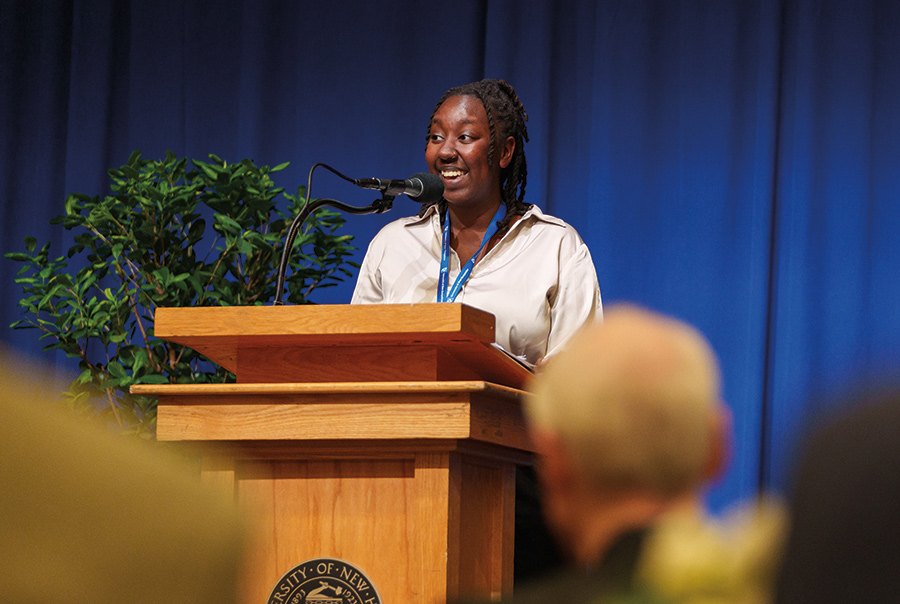 Landscape photograph of Maryrose W. Wainaina ’25 smiling in a light grey/tan blouse and blue lanyard around her neck (dual major in political science and international affairs and executive coordinator of The Black Student Union) as she is speaking at a podium in front of people at the All Hail event in May, which celebrates the stories of possibility at UNH that are created by donor philanthropy.