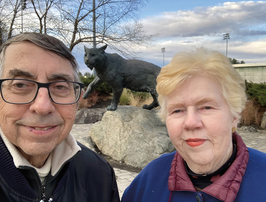 Eric (in a black jacket with prescription see through black outer frame glasses) and Betty Beaverstock (in a blue/maroon jacket wearing red lipstick), both class of 1966, in front of the Wildcat statue outside the Whitt on a recent visit to campus.