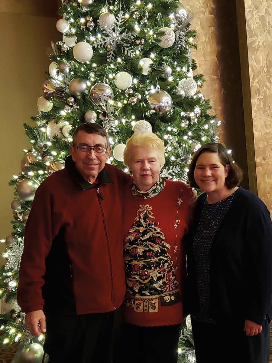 Eric (in a dark red jacket with prescription see through black outer frame glasses) and Betty Beaverstock (in a red Christmas themed cardigan top) smile and pose next to daughter Holly '95 in front of the decorative, beautiful Foxwoods Christmas tree.
