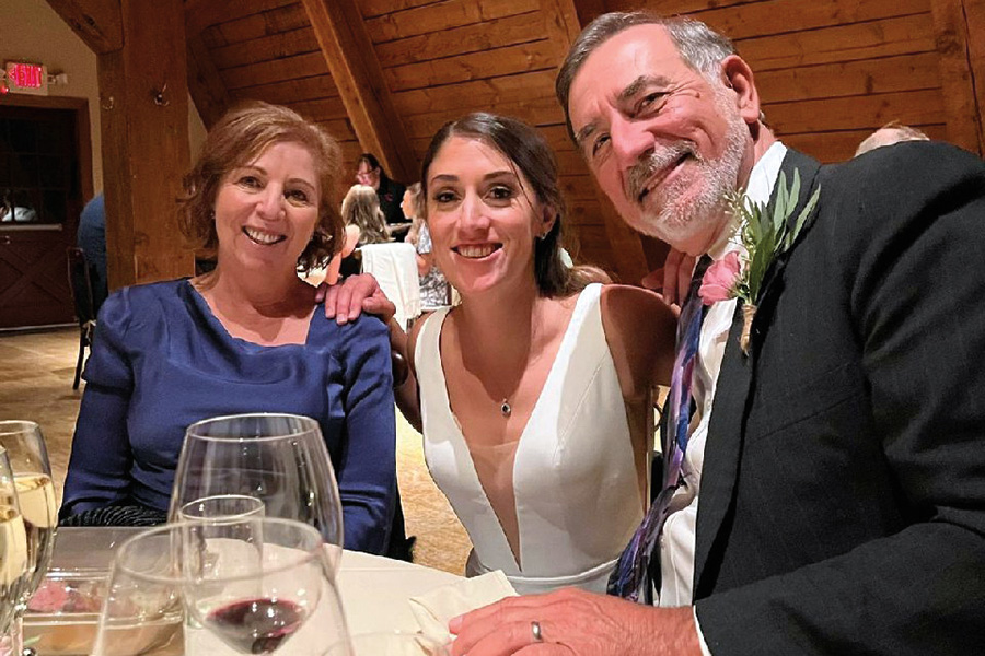 Bernard Roy ’77 (in a black suit and violet space themed tie) and wife Kim (in a dark blue dress) celebrate one of their daughter’s wedding as they all smile and pose next to each other while sitting at a table