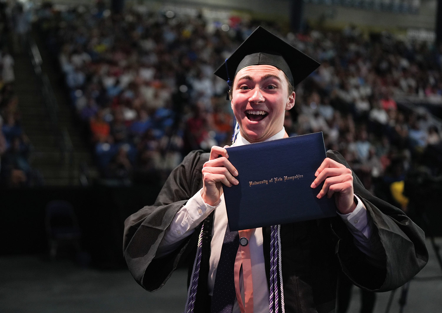 a young man wears a spirited smile, dressed in cap and gown and holding his degree certificate folder up to the camera during the 2023 commencement