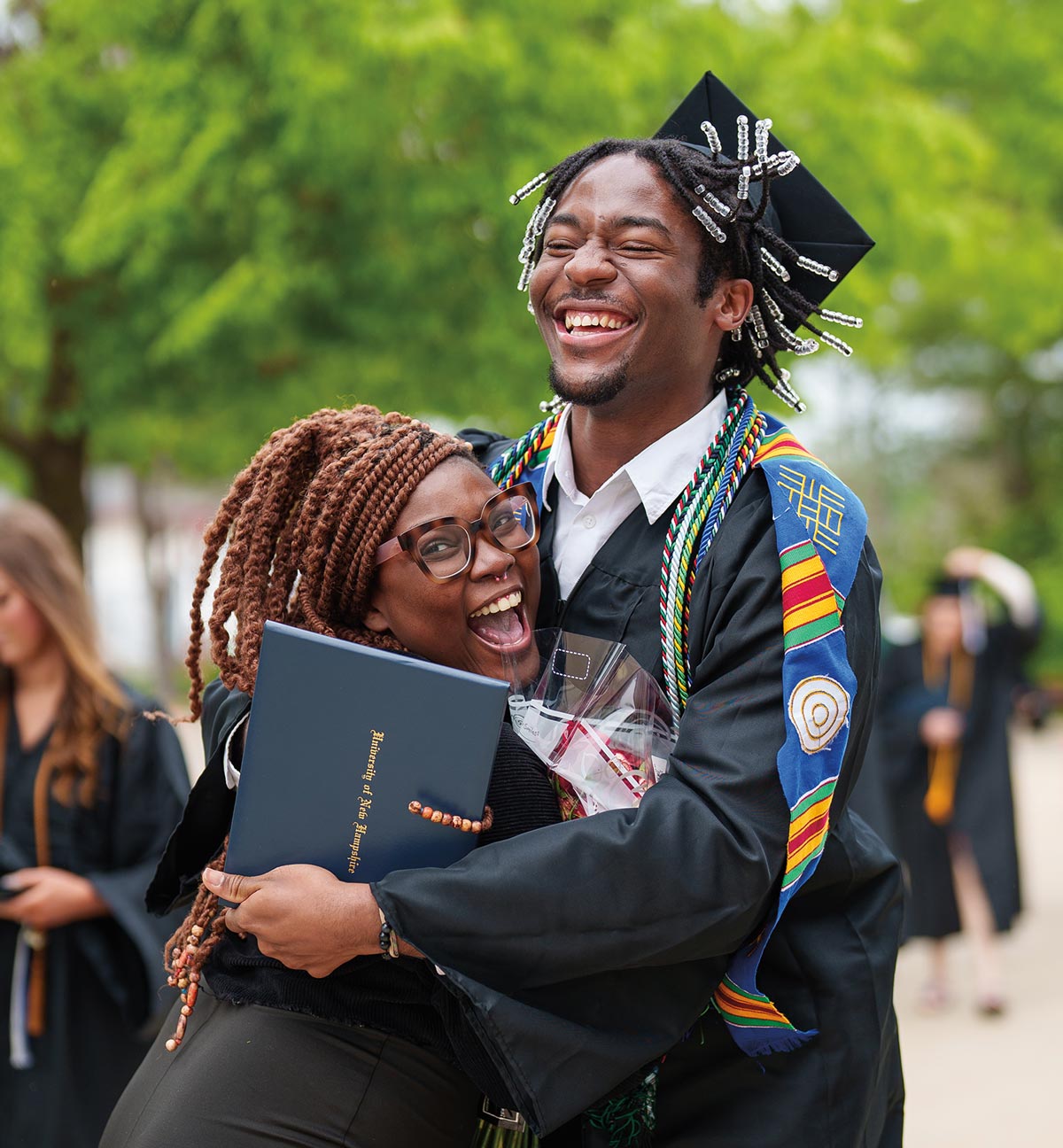 a young man with beaded braids and wearing a cap and gown smiles in mid jump as he hugs a spectacled woman with twisted hair who also wears a wide smile