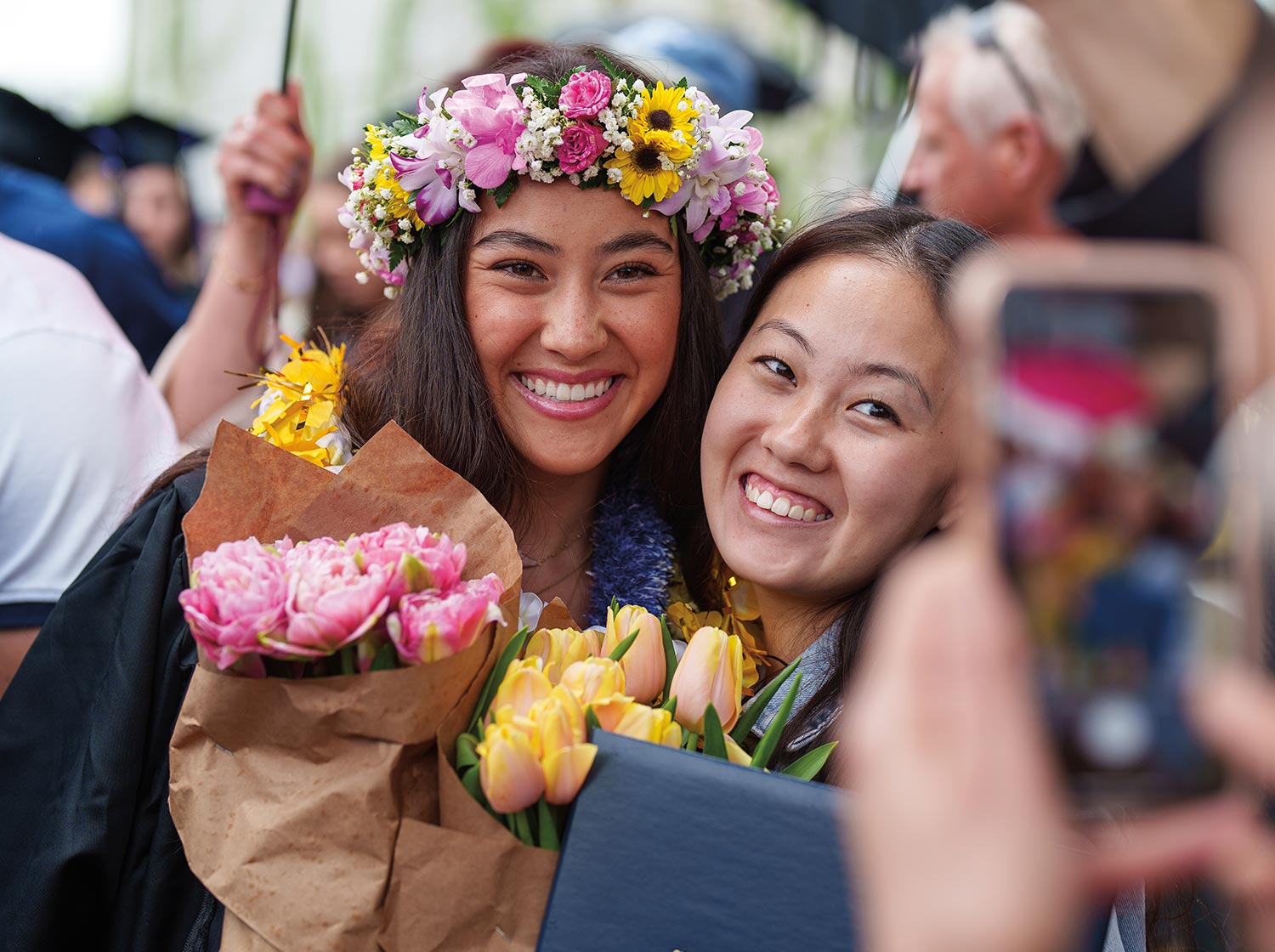 a young woman wearing a gown and a crown of pink and yellow flowers and holding multiple bouquets smiles while taking a photo with another smiling woman