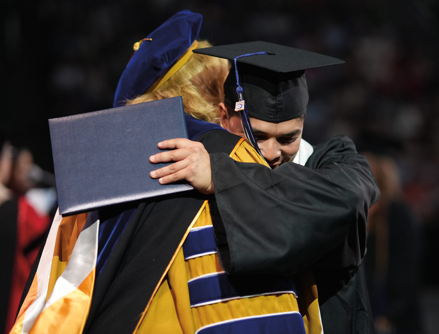 a graduate in a cap and gown holds his certificate in one hand as he hugs and uses the other to shake hands with a faculty member dressed in full regalia