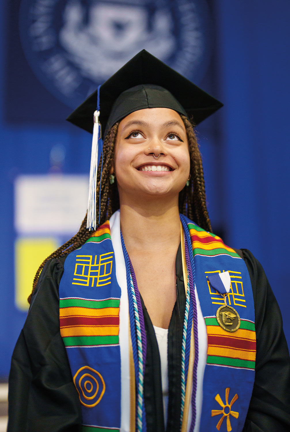 close view of a young woman with with braids wearing a cap, gown and a multi-colored stole smiling as she looks up toward the sky