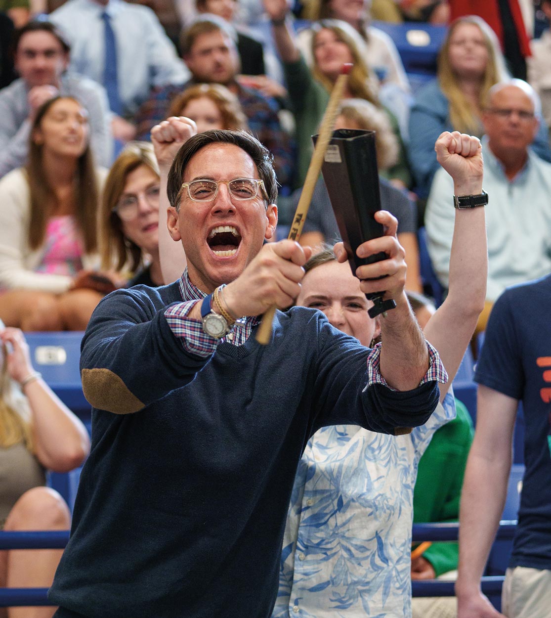 a man photographed in mid yell holds a large cowbell noise maker and a stick among a cheering audience