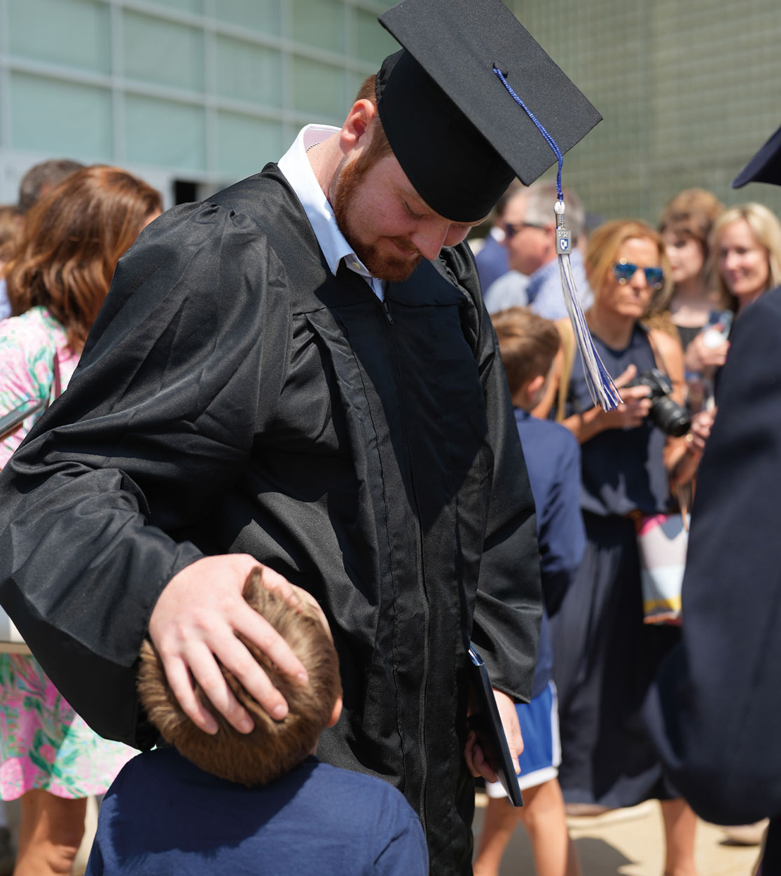 a tall man in a cap and gown looks down playfully at a young boy looking up at the man who rests his hand on the boys head