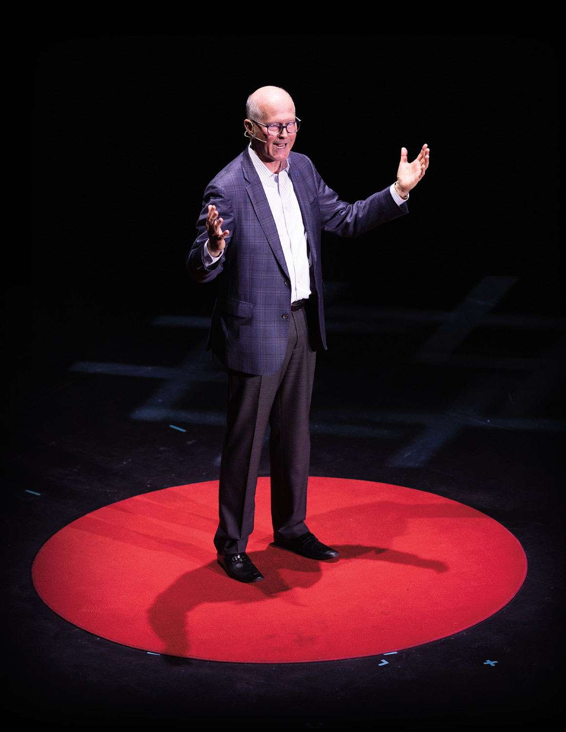 President James W. Dean Jr. delivering his TEDxPortsmouth talk standing on a vibrant red circular carpet in the center of a dark stage