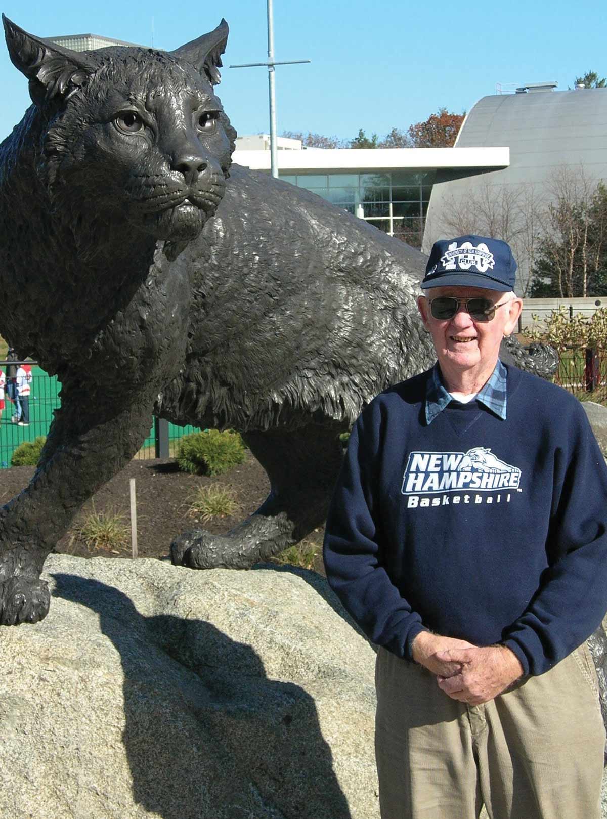 Older man in sweatshirt and ball cap in front of a large statue of a bobcat