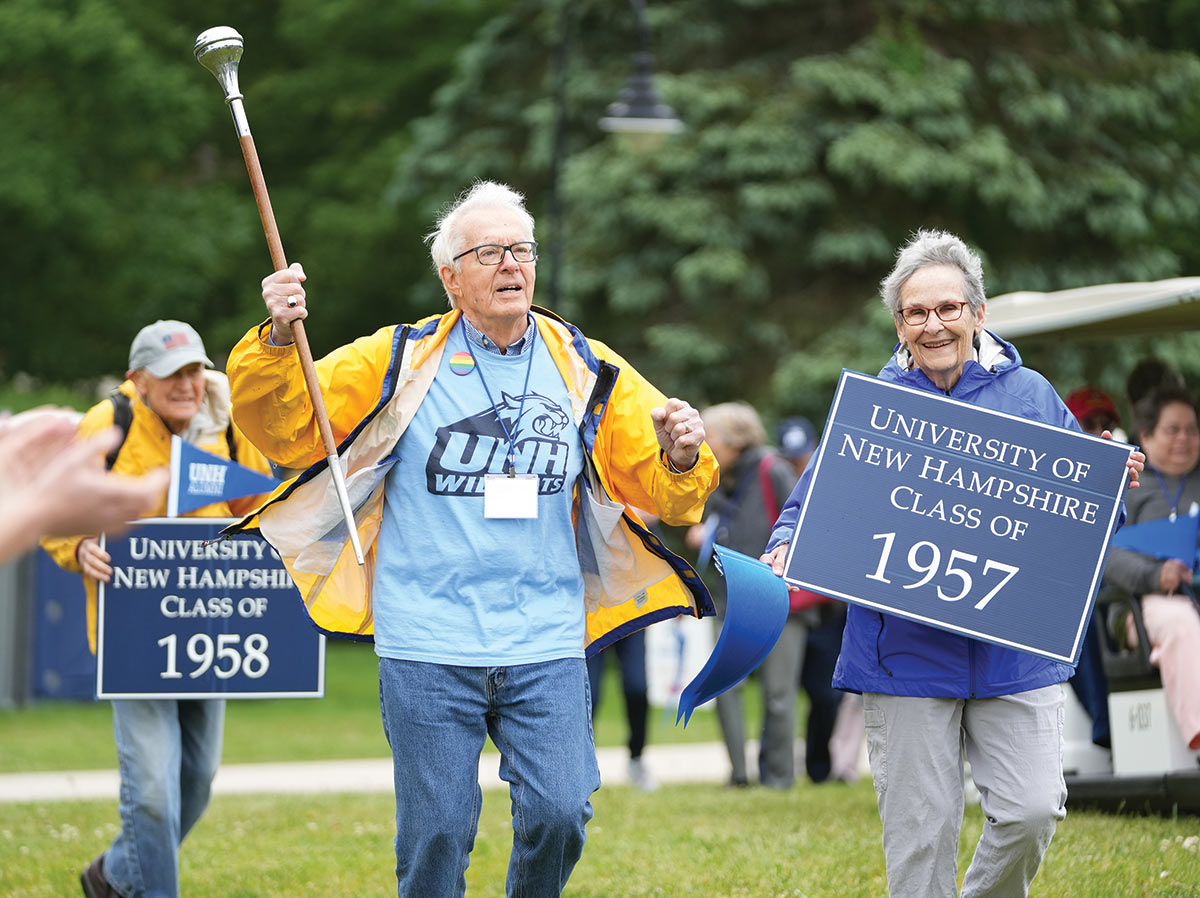 Older man in yellow coat, holding a staff, and an older woman holding a sign that says, University of New Hampshire Class of 1957