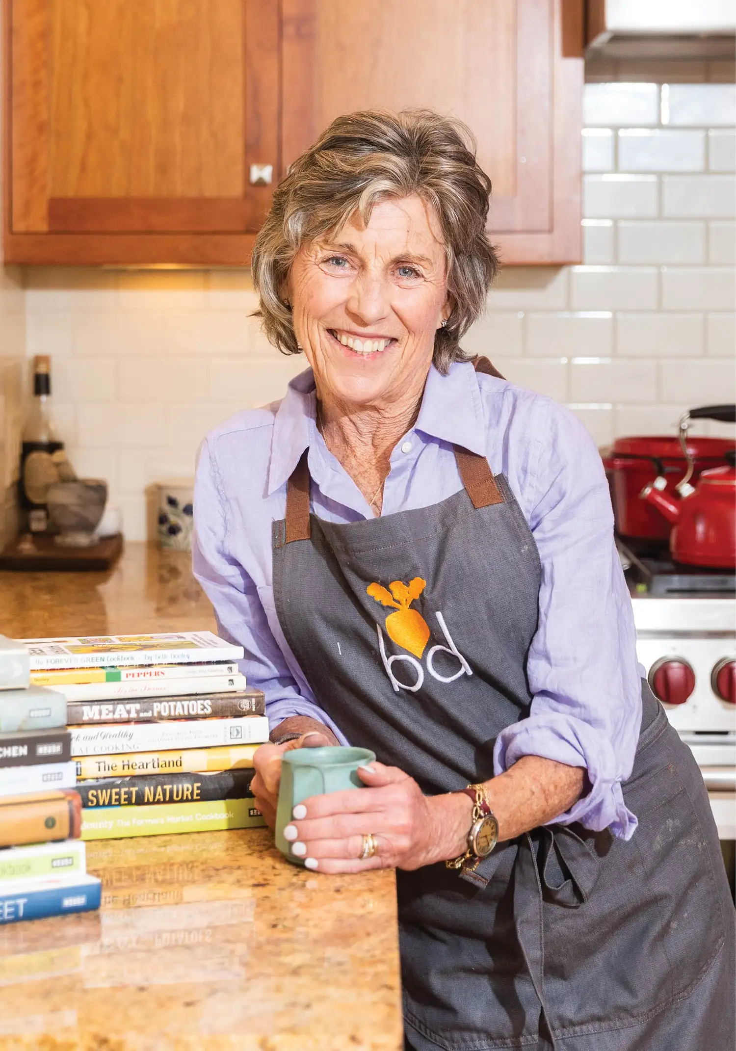 Beth Dooley posing in kitchen with mug, books on counter and wearing apron
