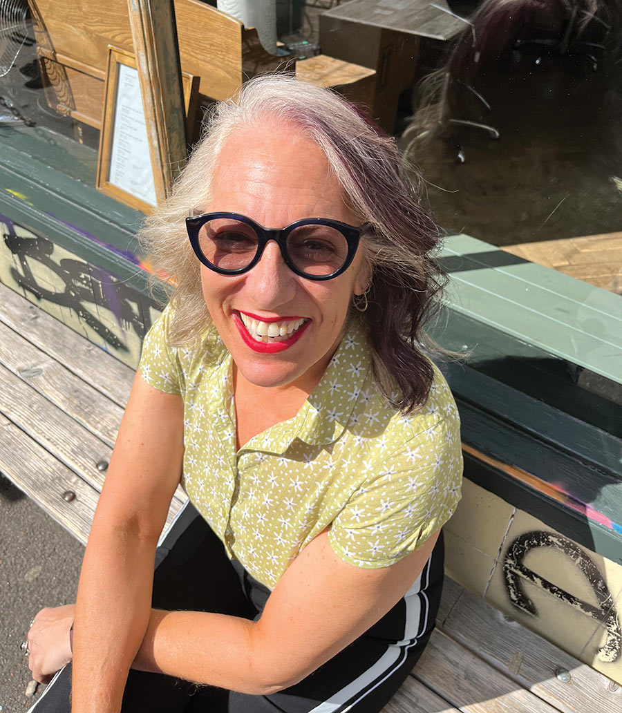 Close-up portrait photo of Suzanne Noble ’83 smiling in a lime green button-up summer shirt and red lipstick while also wearing black outer/inner frame see through prescription glasses as she is seated on a wooden bench outside somewhere