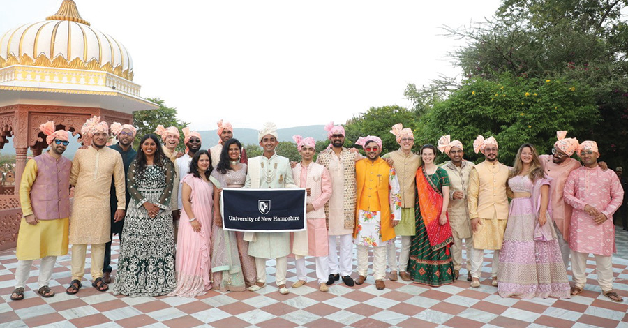 Landscape photograph view of Sig Nigam ’16 and Mimansa Thakar ’17 celebrating their wedding in India in 2023 outside as they pose for a group photo and hold a dark colored small UNH logo banner flag