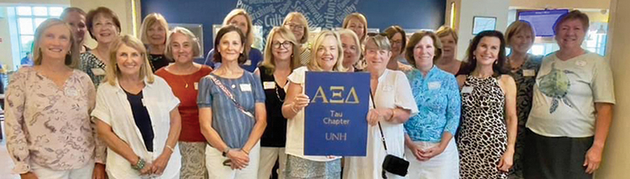 Landscape photograph of a group of Alpha Xi Delta grads from the 1970s who gathered on campus for a reunion with this photo shared by Purr Gow Whalley ’76 as they all are holding a Alpha Xi Delta small ribbon logo banner