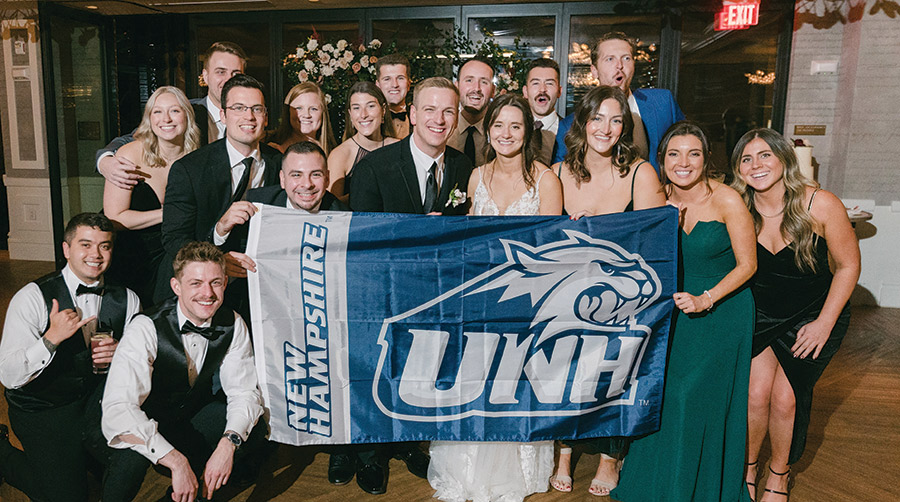 Landscape group photograph of Keegan Smith ’17 and Kathryn Decker ’17 married in December in New Hope, Pennsylvania. Several UNH alumni were in attendance as they all are smiling in joy holding the school-colored UNH logo branded flag indoors somewhere