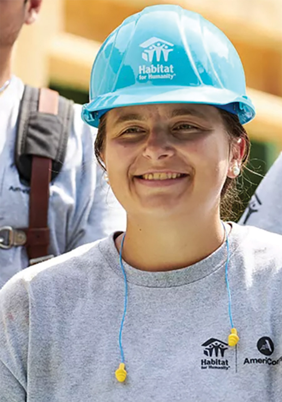 Close-up portrait headshot photograph of Katie Holden ’21 smiling in a light grey Habitat for Humanity t-shirt, has blue/yellow colored ear plug cables around her neck, and is wearing a turquoise Habit for Humanity logo branded hardhat standing outdoors somewhere
