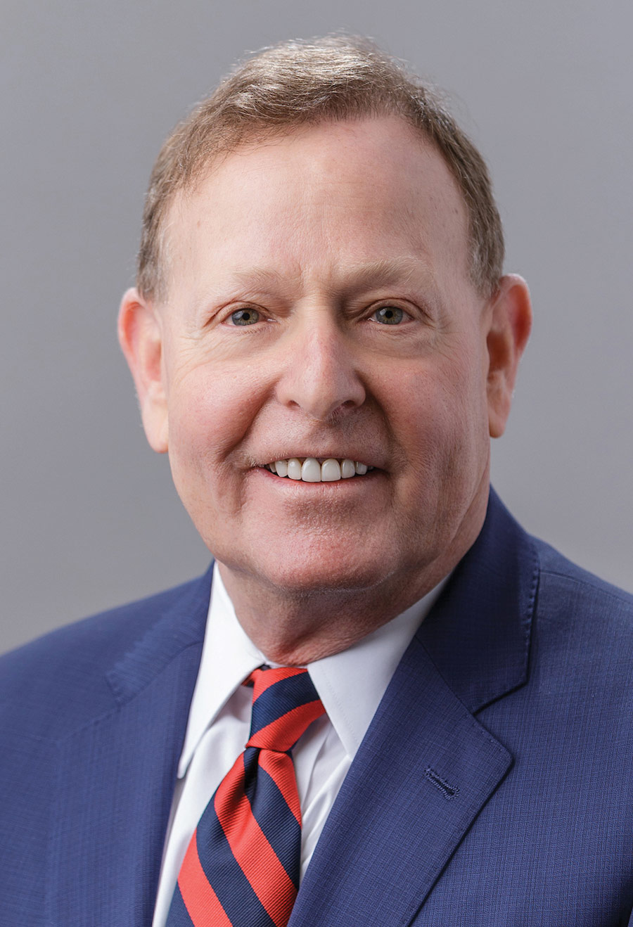 Portrait headshot photograph of Dr. Robert Barish smiling in a dark navy blue blazer suit and white button-up dress shirt underneath with a multi-colored pattern design tie (red/dark navy blue)