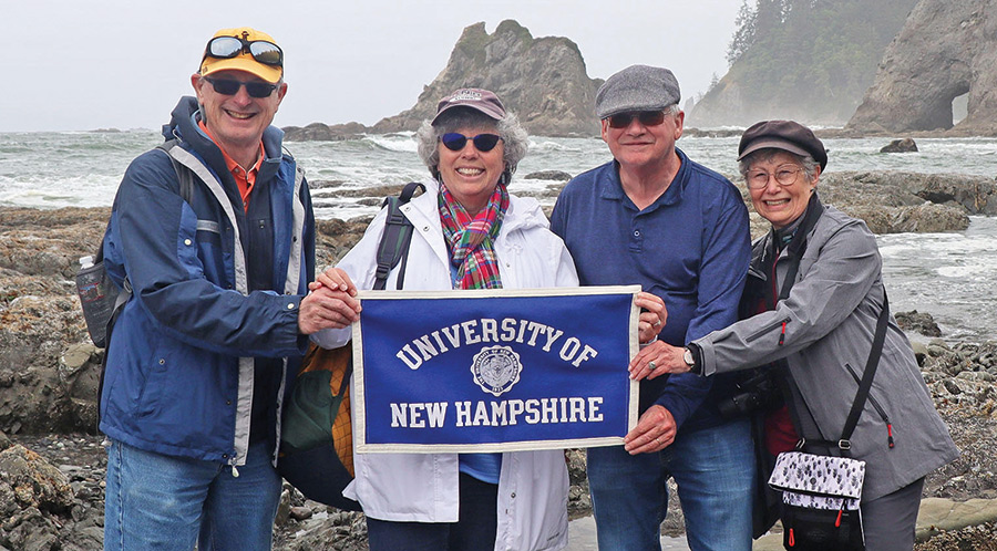 Landscape photograph of (Left to right, Charles “Kirk” Taft, Jr. ’76 and Nancy Waldman Taft ’77; Daniel D. Stanton (Kirk’s UNH roommate) ’76; and Pamela Hamilton-Powell ’76. Dan’s wife, Clara Conner, is not pictured) as all of them are holding a small blue UNH logo seal banner flag out on the rocks near the ocean somewhere
