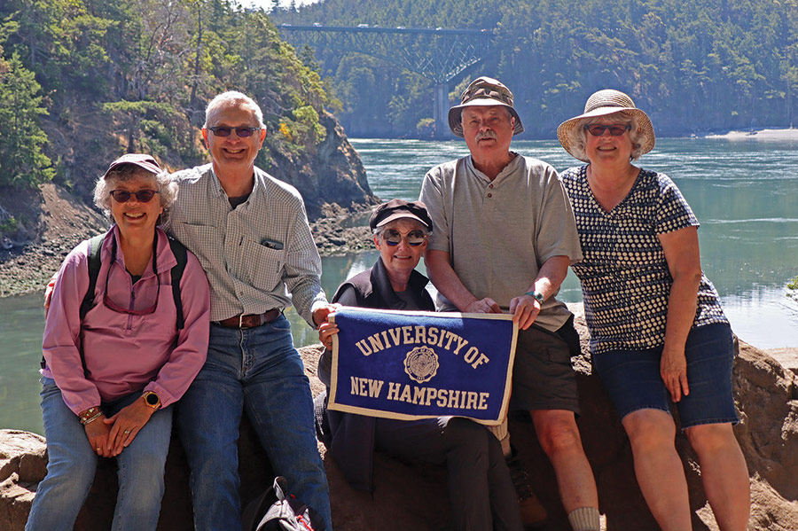 Landscape photograph of (Left to right: Nancy Waldman Taft ’77 and Charles “Kirk” Taft, Jr. ’76 of Gig Harbor, Washington; Pamela Hamilton-Powell ’76 of Atlantic Beach, Florida; Jim Fels ’75 and Linda Doherty Fels ’77 of Bellingham, Washington. Kirk, Pam, and Jim are all retired from careers in the U.S. Air Force. Pam also retired from the Federal Aviation Administration) as all of them are posing for a group photo together holding a blue UNH seal logo banner flag outside somewhere