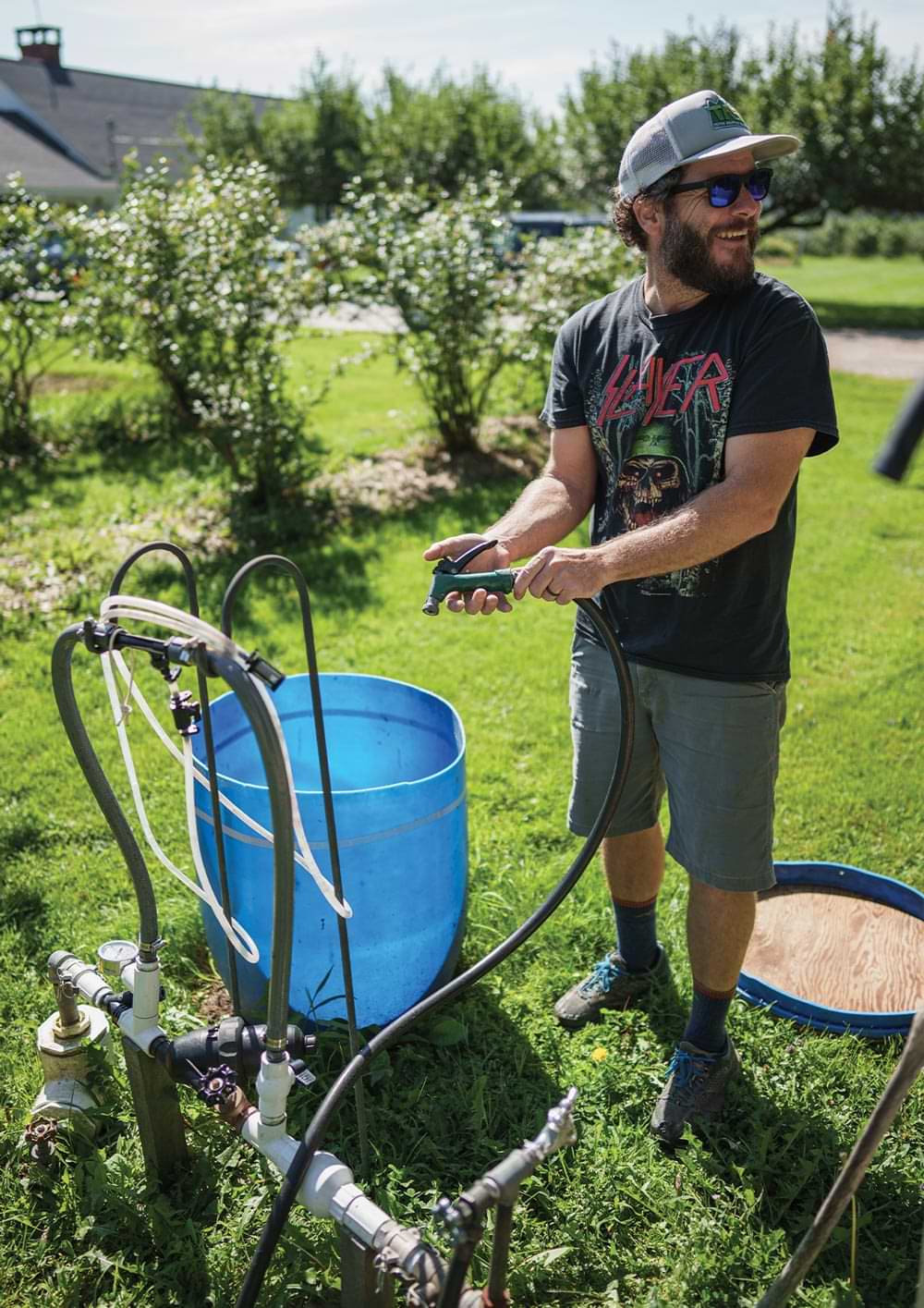 Pete Bartlett, wearing a cap, a t-shirt, shorts, and sunglasses, looks off camera while holding a water hose and standing next to a large plastic blue barrel on a vibrant green yard
