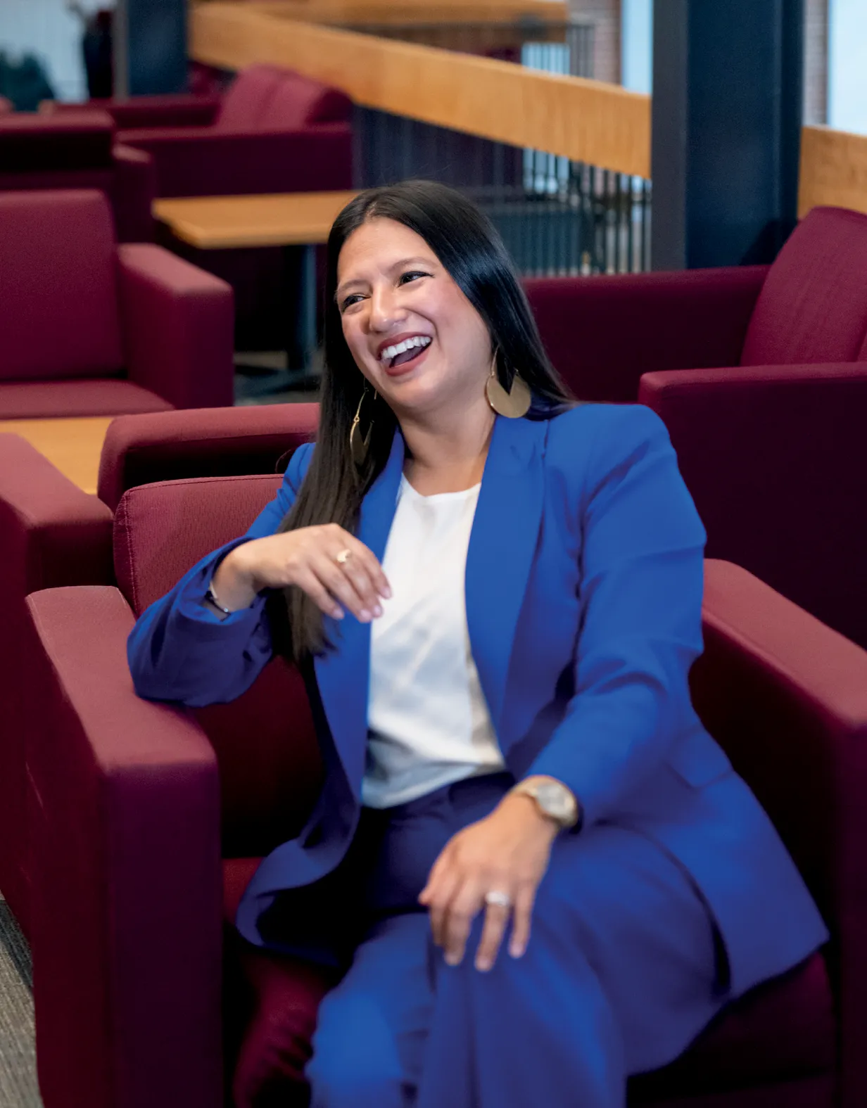 A woman with long dark hair, wearing a royal blue suit and a white blouse, sits on a maroon armchair in a modern lounge area. She is laughing and gesturing with her hand.