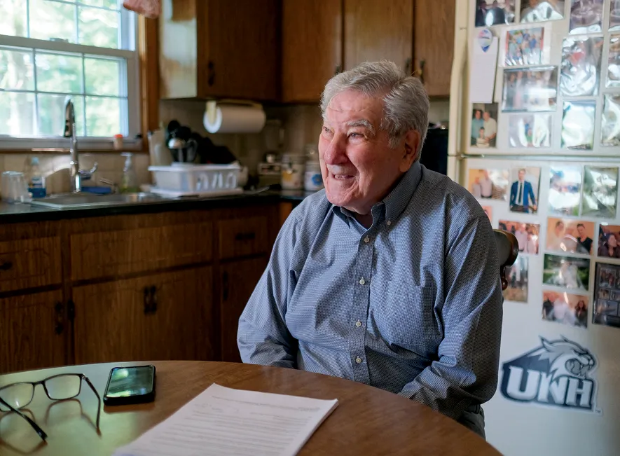 An elderly man with gray hair, wearing a blue button-down shirt, sits at a wooden table in a home kitchen. A paper document, glasses, and a smartphone are on the table. Behind him, a refrigerator is covered with family photos, and a window provides natural light.