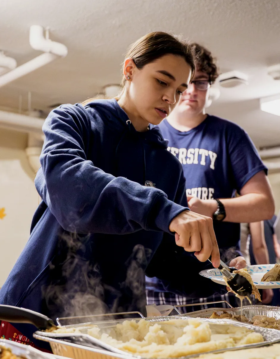 A young woman in a navy hoodie serves food onto a paper plate in a cafeteria or dining hall. A young man in a University of New Hampshire t-shirt stands behind her holding a plate.