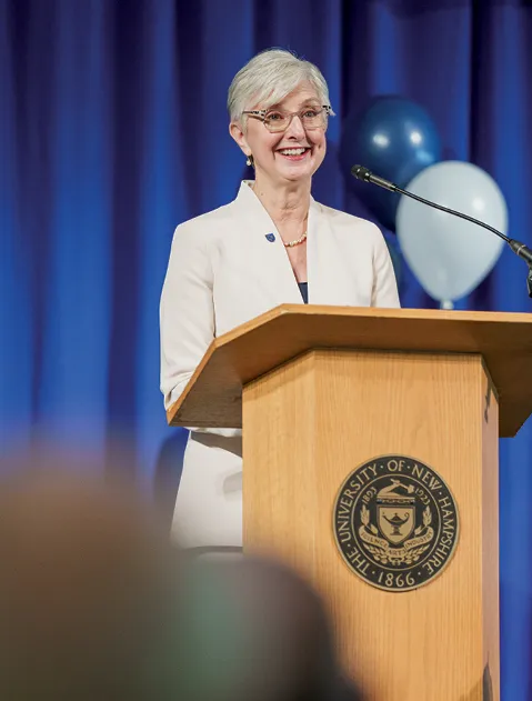 A woman with short gray hair and glasses, wearing a cream-colored blazer, stands at a wooden podium with the University of New Hampshire seal. She smiles while speaking into a microphone. Blue curtains and balloons are in the background.