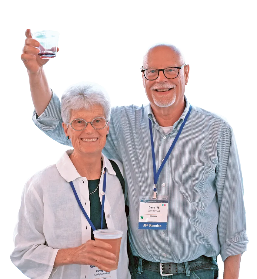 Older couple at a reunion event smiling at the camera, each holding a drink, with name tags and lanyards indicating “Class of ’75 50th Reunion.”