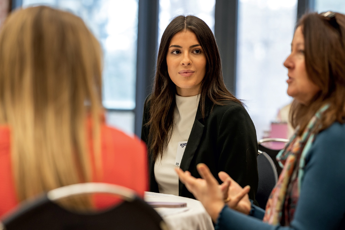 brunette female with white blouse and black blazer looking at other females that are out of focus in front of her at a table