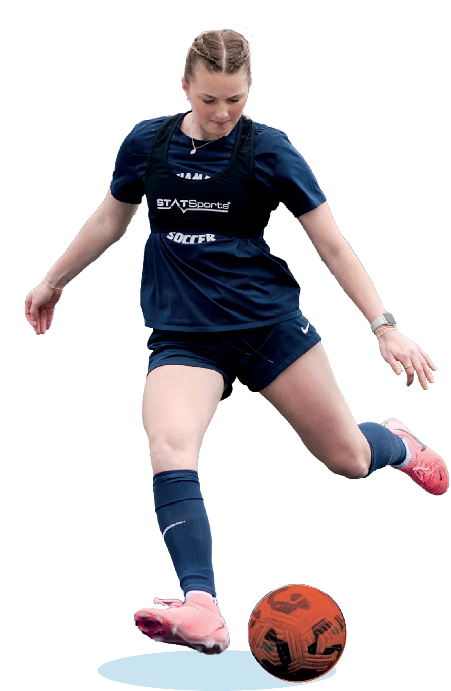 Female soccer player in navy blue uniform and pink cleats kicking an orange ball while wearing a STATSports performance vest.