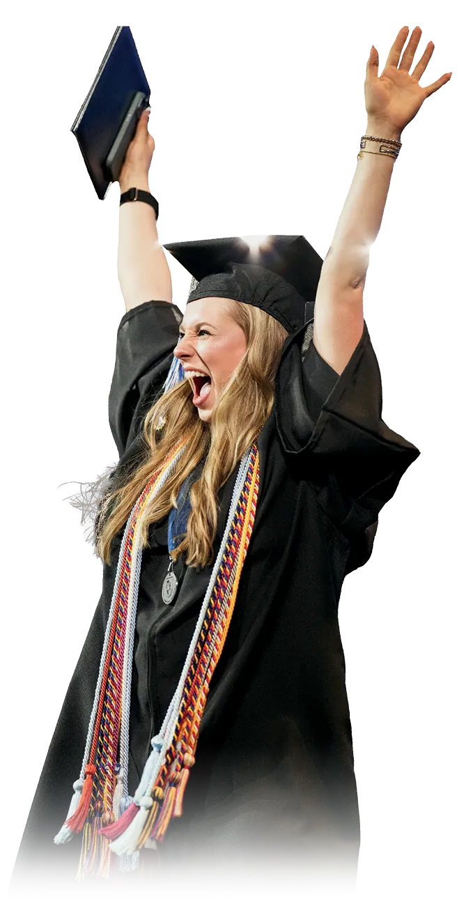 Excited graduate in cap and gown raising both arms and holding a diploma, wearing multiple colorful honor cords and a medallion.