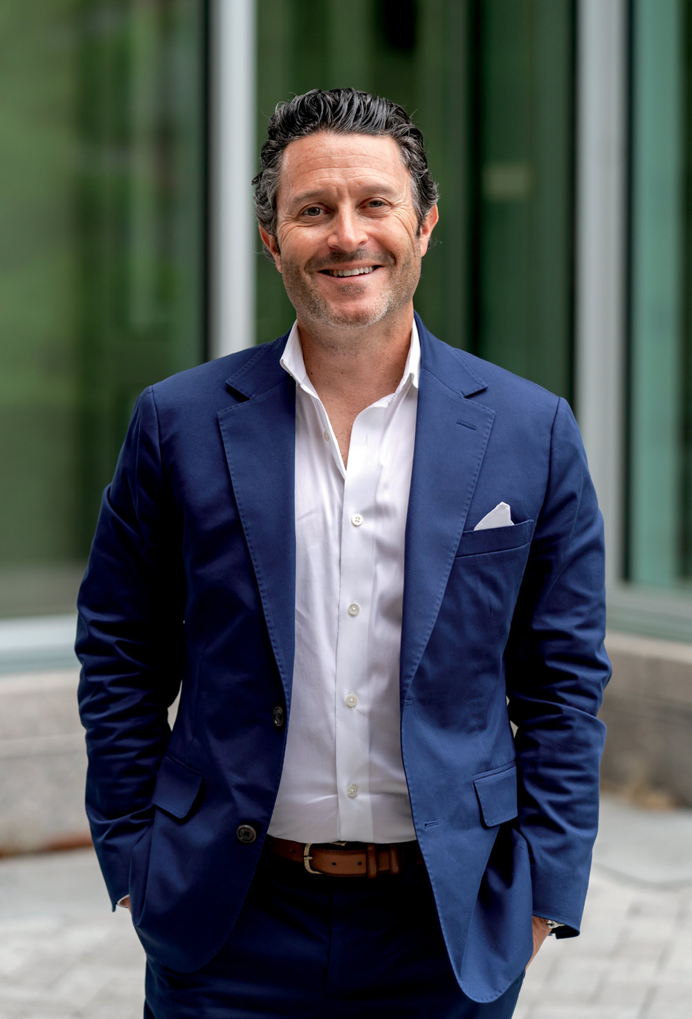 Person in a blue suit standing in front of a glass building, smiling.