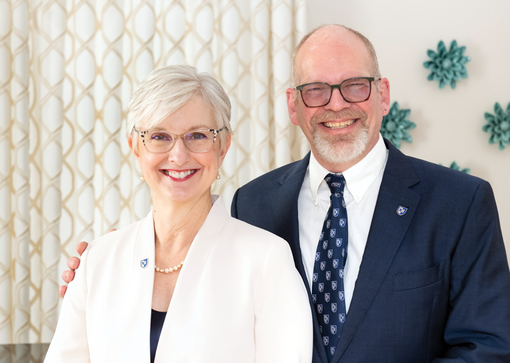 Two people smiling, one in a light blazer, the other in a dark suit, standing in front of a patterned curtain and decorative wall pieces.